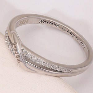 Może przedstawiać: Srebrny pierścionek z krzyżowym wzorem i małymi, przezroczystymi kamieniami. Na pierścionku wygrawerowano słowa "Good Girl" i "Nothing is impossible".