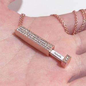 May include: A rose gold necklace with a rectangular pendant that is covered in clear rhinestones. The pendant opens to reveal a small metal plate with the inscription "HotWife".