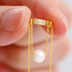 Puede incluir: Un delicado collar de oro con un pequeño colgante de perla redonda. El collar tiene una fina cadena y una barra rectangular en la parte superior con la inscripción "Good Girl". La joya se sostiene sobre un fondo suave y borroso.