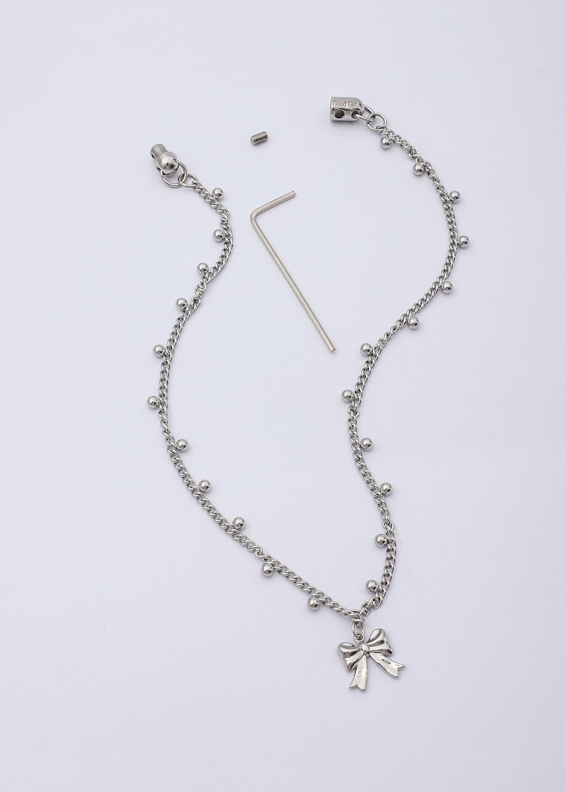 May include: Silver-toned necklace with a bow pendant. The necklace features a delicate chain design with small, evenly spaced ball accents. Includes a small screw and a silver-toned hex key. The bow pendant adds a touch of elegance.