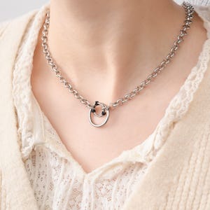 Puede incluir: Collar de cadena plateada con un colgante circular. El collar presenta una cadena gruesa y redondeada y un pequeño colgante de anillo con un acento negro. La joya se lleva sobre una parte superior de color crema.