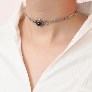 Peut inclure: Un collier ras du cou argenté avec un petit pendentif en perle noire. Le collier a un design de chaîne délicat avec un pendentif circulaire tenant la perle. Le collier est porté contre une chemise blanche.