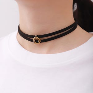 Puede incluir: Un collar gargantilla negro con dos bandas y un colgante circular de color dorado. El collar se lleva alrededor del cuello. La persona lleva una camiseta blanca.