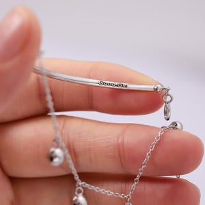 May include: A silver-toned bracelet with the inscription "Good Girl" and three dangling spherical charms. The bracelet is held in a hand against a soft, neutral background, highlighting its delicate design.