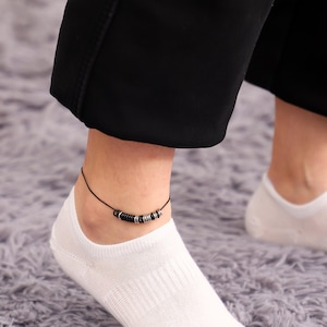 May include: A black and silver beaded ankle bracelet on a person's ankle wearing white socks.