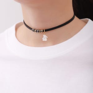 Puede incluir: Gargantilla negra con detalles dorados y plateados y un pequeño colgante de conejo blanco. El collar se lleva alrededor del cuello. La persona lleva una camiseta blanca. El texto "thinkAndKink.com" es visible en la camiseta.