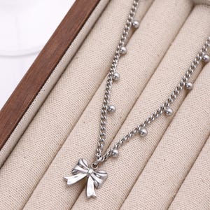May include: A silver-colored necklace with a bow pendant. The necklace features a chain design with small, round embellishments. The bow pendant is detailed and sits at the center. The necklace is displayed in a wooden jewelry box with a beige fabric lining.