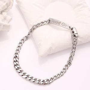 May include: A silver-colored chain bracelet with a rectangular plate that reads "Good Girl." The bracelet has a curb chain design and a clasp closure. The jewelry is displayed on a white surface.