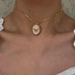 Könnte beinhalten: Eine goldene Kettenkette mit einem weißen ovalen Anhänger mit einem goldenen Schmetterlingsdesign.
