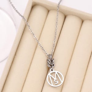 May include: A silver-colored necklace with a delicate chain and a pendant. The pendant features a circular design with the letters "M" and "S" inside. The necklace is displayed on a beige surface.