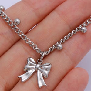 May include: Silver-toned chain bracelet with a bow pendant. The bracelet features a delicate chain design with small, round beads interspersed along the chain. The bow pendant is a shiny, three-dimensional design.