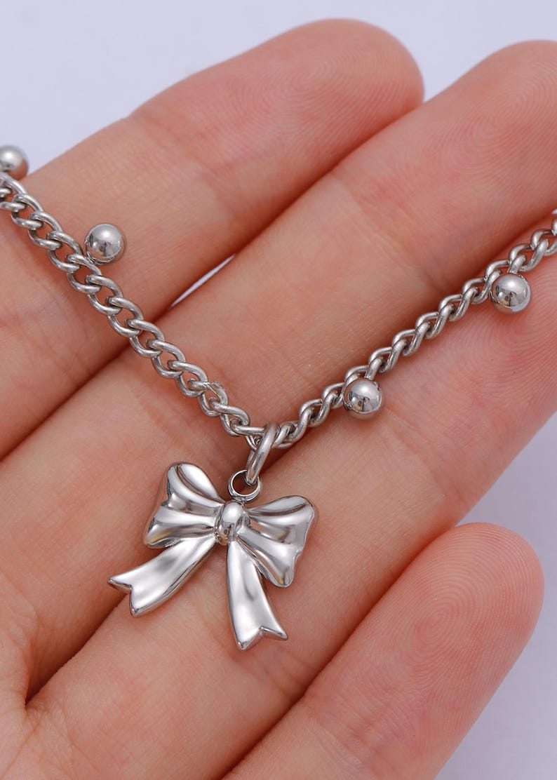 May include: Silver-toned chain bracelet with a bow pendant. The bracelet features a delicate chain design with small, round beads interspersed along the chain. The bow pendant is a shiny, three-dimensional design.