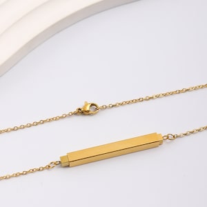 Könnte beinhalten: Eine goldene Kettenkette mit einem langen, rechteckigen Anhänger. Der Anhänger ist aus glattem, glänzendem Metall.