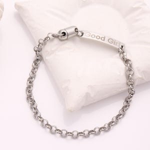 May include: A silver-colored bracelet with a chain-link design. The bracelet features a rectangular bar with the engraved text "Good Girl". The bracelet is made of a shiny, metallic material and is displayed on a white surface.