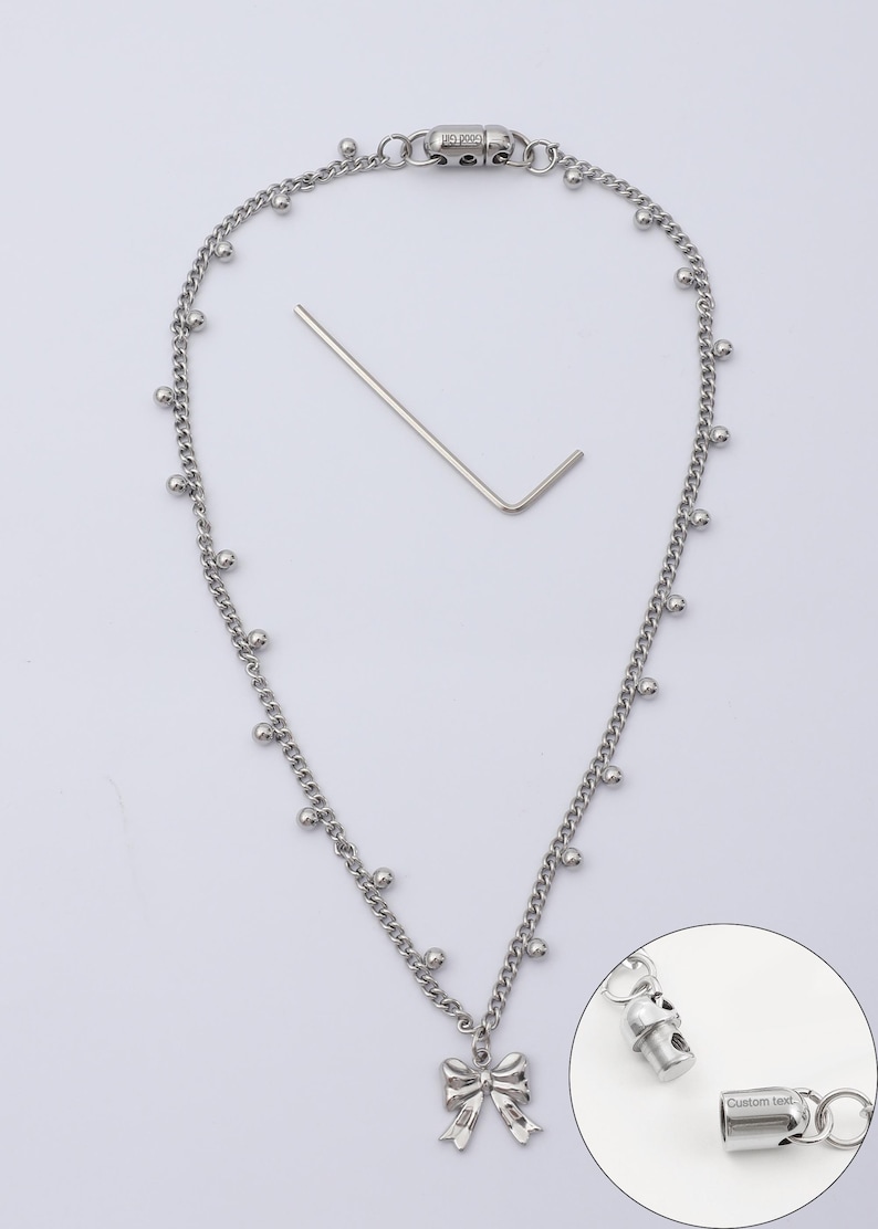 May include: Silver-toned necklace with a bow pendant and small ball accents. Includes a small metal tool and a close-up of the clasp with the text "Custom text". The necklace is on a white background.