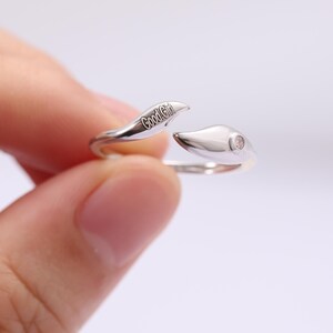 Puede incluir: Un anillo plateado con un diseño abierto. Un extremo del anillo está grabado con el texto "Good Girl", y el otro extremo presenta una pequeña piedra redonda y transparente. El anillo se muestra sobre un fondo claro.