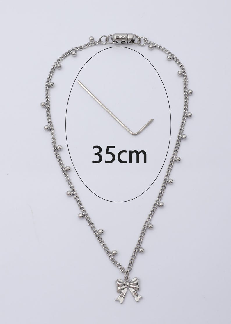 May include: Silver-toned necklace with a bow pendant. The necklace is 35cm (13.8 inches) long and features small ball accents along the chain. A small wrench is included. The necklace has a clasp closure.