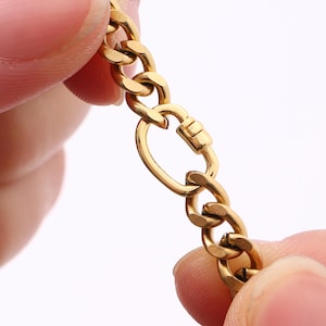 May include: Close-up of a gold-coloured chain necklace with a lobster clasp. The chain links are thick and rounded. The clasp is attached to a large oval link. The necklace is held by two hands.