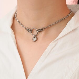 Peut inclure: Collier ras du cou argenté avec un pendentif en forme de cœur. Le collier a un design de chaîne avec une pièce maîtresse circulaire. Le pendentif en forme de cœur est suspendu au centre. Le collier est porté par une personne portant une chemise blanche.