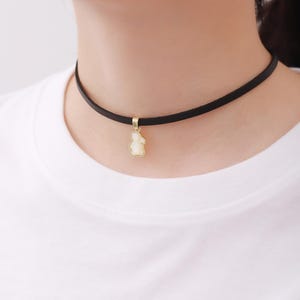 Peut inclure: Un collier ras du cou noir avec un pendentif doré en forme d'ours. Le pendentif a un centre blanc nacré. Le collier est porté sur un t-shirt blanc.