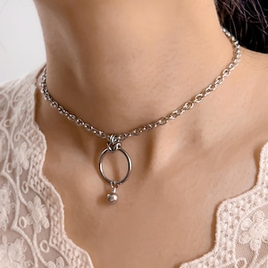 Op de afbeelding: Een zilverkleurige choker ketting met een kettingontwerp. De ketting heeft een grote cirkelvormige hanger met een kleine, ronde bedel. De ketting wordt om de nek gedragen.