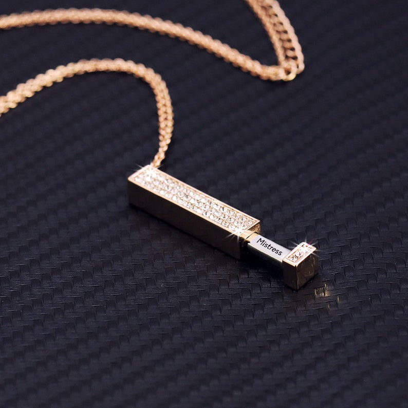 May include: A gold chain necklace with a rectangular pendant encrusted with clear rhinestones. The pendant opens to reveal a small compartment with the word "Mistress" engraved inside.