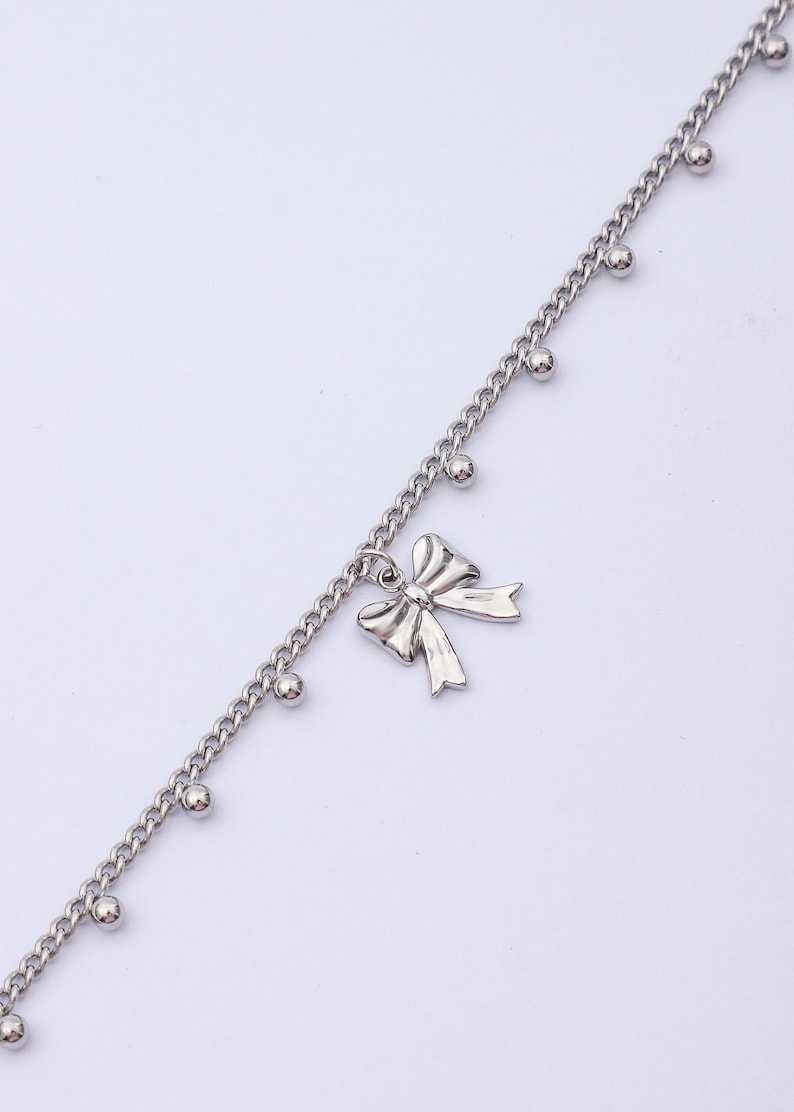 May include: Silver-toned anklet featuring a delicate bow pendant. The chain is adorned with small, round beads. This piece of jewelry is designed to be worn around the ankle.
