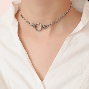 Può includere: Una collana girocollo color argento con un design a catena e un ciondolo circolare. La collana è indossata intorno al collo. Lo sfondo è rosa tenue.
