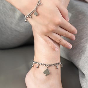 Könnte beinhalten: Silberfarbene Fußkettchen mit Kettenmuster. Jedes Fußkettchen hat einen Pik-Anhänger und kleine, baumelnde Anhänger. Die Fußkettchen werden am Knöchel und Fuß getragen.