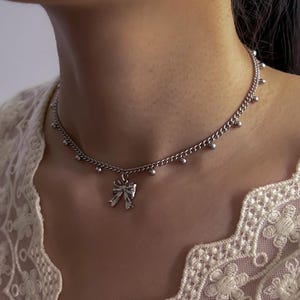May include: Silver-toned necklace featuring a delicate bow pendant. The chain is adorned with small, round beads. The necklace is worn against a backdrop of a white lace top, creating a soft, feminine aesthetic. A simple yet elegant piece of jewelry.