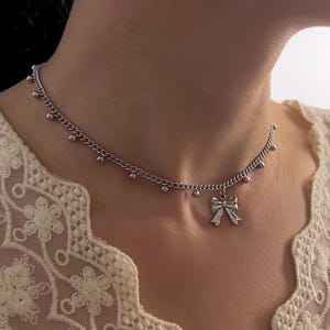 May include: Silver-toned necklace featuring a delicate bow pendant. The chain is adorned with small, evenly spaced beads. The necklace is worn around the neck, showcasing its simple yet elegant design. The background is a cream-colored lace.