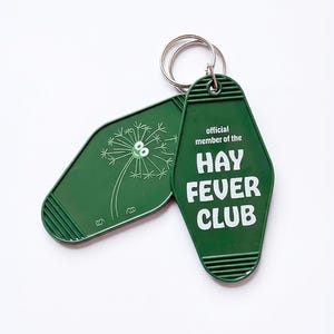 Op de afbeelding: Twee groene sleutelhangers met zilveren sleutelringen. Eén sleutelhanger heeft een paardenbloemillustratie, de andere leest "official member of the HAY FEVER CLUB" in witte tekst. De sleutelhangers zijn ruitvormig.