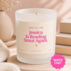 May include: A frosted glass candle in a cylindrical container with a cream-colored label. The label features pink text reading "SMELLS LIKE Jessica Is Reading Smut Again." and "CHERRY ROSE COLLECTION," with "BURN TIME: 39 HRS" and "SOY WAX" printed.