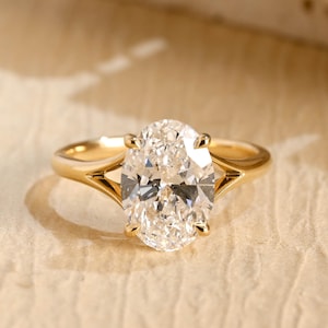 Puede incluir: Un anillo de oro con una gran piedra preciosa transparente de talla ovalada y facetada, montada en un engaste de cuatro garras. La banda presenta un diseño de brazo dividido que se estrecha hacia la piedra central, con un acabado metálico pulido.