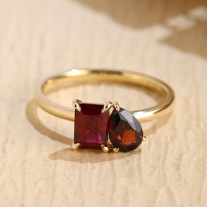 Pode incluir: Um anel de ouro com duas pedras preciosas distintas colocadas lado a lado. Uma pedra é uma gema vermelha profunda de corte esmeralda retangular, enquanto a outra é uma pedra menor de cor vermelho-escura em formato de gota.
