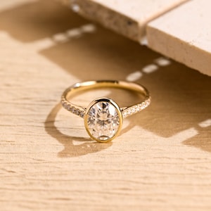 May include: A gold engagement ring with a large oval-shaped diamond set in a bezel setting. The band is adorned with a row of smaller diamonds.