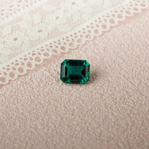 May include: A deep green emerald gemstone with a rectangular cut, set against a pink textured background.