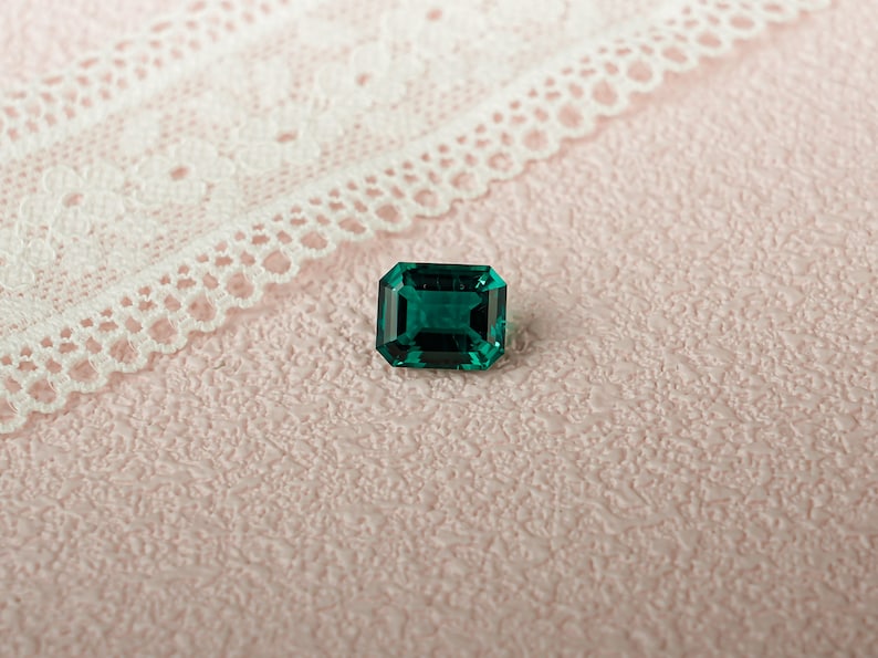 May include: A deep green emerald gemstone with a rectangular cut, set against a pink textured background.