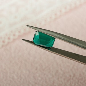 May include: A vibrant green gemstone held by tweezers. The gemstone is a square shape with beveled edges and a smooth, polished surface.