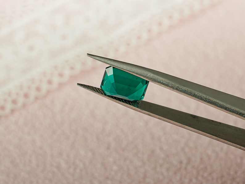 May include: A vibrant green gemstone held by tweezers. The gemstone is a square shape with beveled edges and a smooth, polished surface.