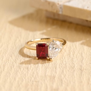 Può includere: Un anello d'oro con una gemma rettangolare rosso intenso e una gemma trasparente a forma di pera. L'anello è posto su una superficie beige chiaro, con ombre morbide e uno sfondo sfocato.