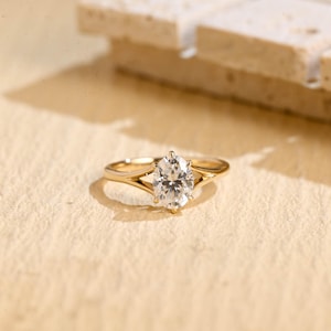 Könnte beinhalten: Ein goldener Ring mit einem großen, oval geformten, klaren Edelstein. Der Ring hat ein geteiltes Schaftdesign, wobei sich das Band in zwei zarte Stränge auf beiden Seiten des Edelsteins aufteilt. Der Ring ist vor einem neutralen Hintergrund platziert.