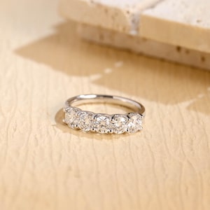 May include: A silver ring featuring five clear, round gemstones. The ring is set on a light tan surface. The background is blurred, showing a light-coloured, textured object.