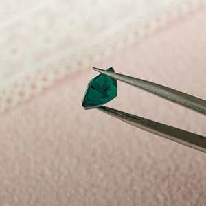 May include: A pair of tweezers holding a dark green gemstone. The gemstone is faceted and has a triangular shape.