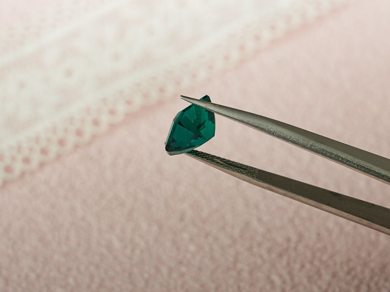 May include: A pair of tweezers holding a dark green gemstone. The gemstone is faceted and has a triangular shape.