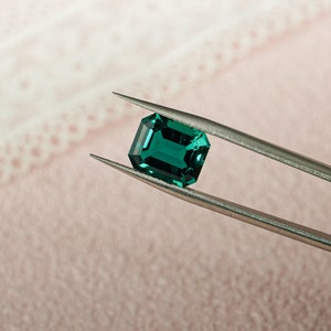 May include: A vibrant green emerald gemstone held in tweezers. The emerald is a rectangular cut with a smooth, polished surface.