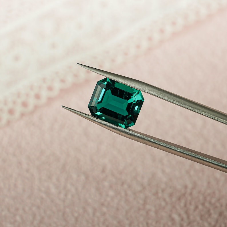 May include: A vibrant green emerald gemstone held in tweezers. The emerald is a rectangular cut with a smooth, polished surface.