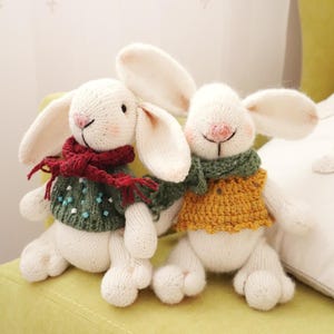May include: Two white knitted bunny stuffed animals with long ears, rosy cheeks, and smiling faces. One bunny wears a green sweater and a red scarf, the other wears a yellow sweater and a green scarf. They are sitting on a green surface.