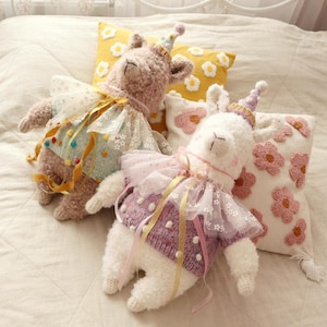 May include: Two handmade llama soft toys wearing party hats and decorative outfits. One is light brown with a blue and yellow outfit, the other white with a purple outfit. Two decorative pillows with floral designs are in the background.
