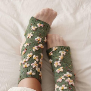 Puede incluir: Calentadores de piernas de punto verde adornados con adornos florales blancos y amarillos. Los calentadores de piernas están decorados con pequeñas cuentas de colores. El fondo es una sábana blanca.